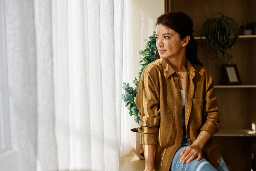 Perfil lateral de una mujer vestida con una camisa marrón, sentada junto a una ventana con cortinas blancas semitransparentes.