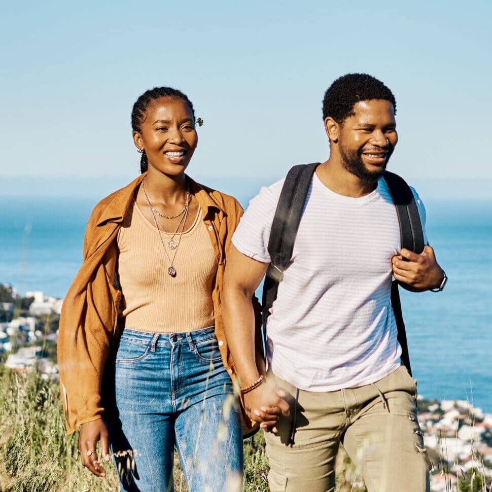 Pareja caminando de la mano por una ladera costera con el océano azul y el cielo detrás de ellos, sonriendo mientras caminan.
